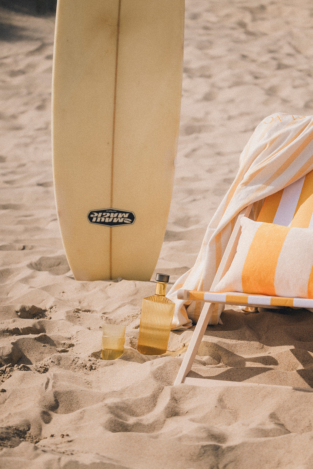 A stylish beach setup with a surfboard, striped deckchair, and an elegant reusable water bottle and cup placed in the sand.